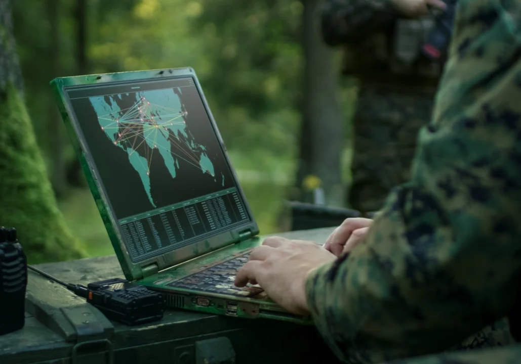 Person in camouflage uses a laptop displaying a world map with data connections. A walkie-talkie is nearby.